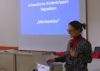 Workshop 3 von Mag. Reisenberger. Sie steht vor ihrem Laptop und lächelt. Im Hintergrund ist ihre Präsentation zu sehen.