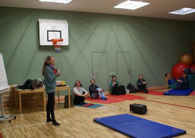 Workshop 5 von Frau Brencic. Sie steht im Gymnastikraum des BBI und spricht mit ihren Workshop-Teilnehmer*innen, welche vor ihr am Boden auf Matten Platz genommen haben.