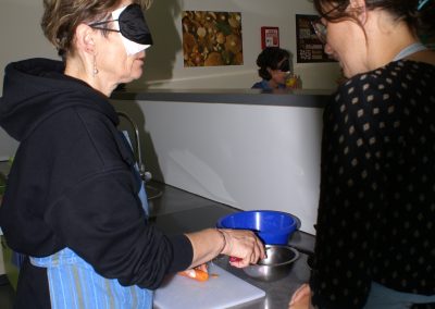 Workshop 10 von Fr. Löbl und Fr. Daniel. Fr. Löbl leitet eine Workshopteilnehmerin, welche eine Simualtionsbrille für Blindheit trägt, an, wie man ohne zu sehen Gemüse mit einem Messer sicher schneidet.